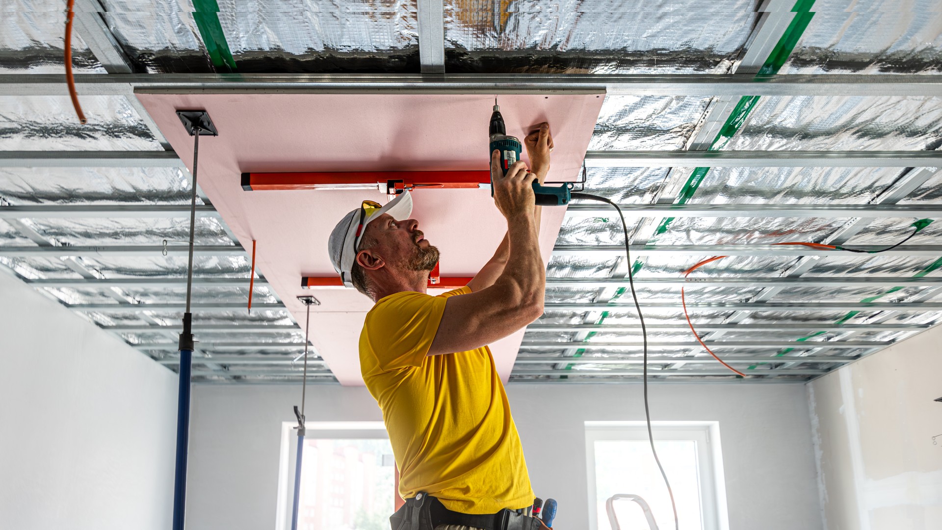 The worker screwing plasterboard to the ceiling. He is using special electric screwdriver. A red plasterboard improve the fire resistance of ceiling structures.